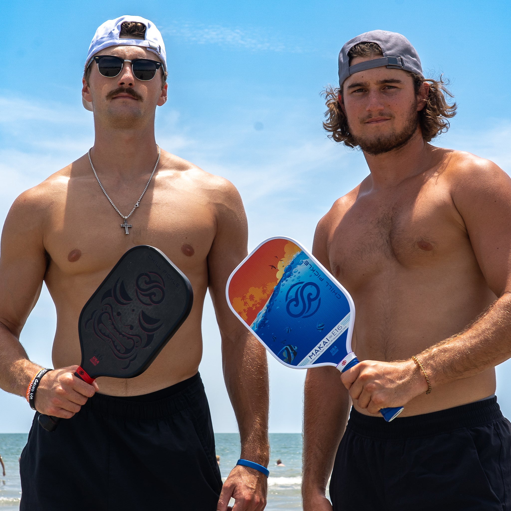 Two men holding Chill Pickle Kahuna pickleball paddles on a sunny beach court in casual playwear