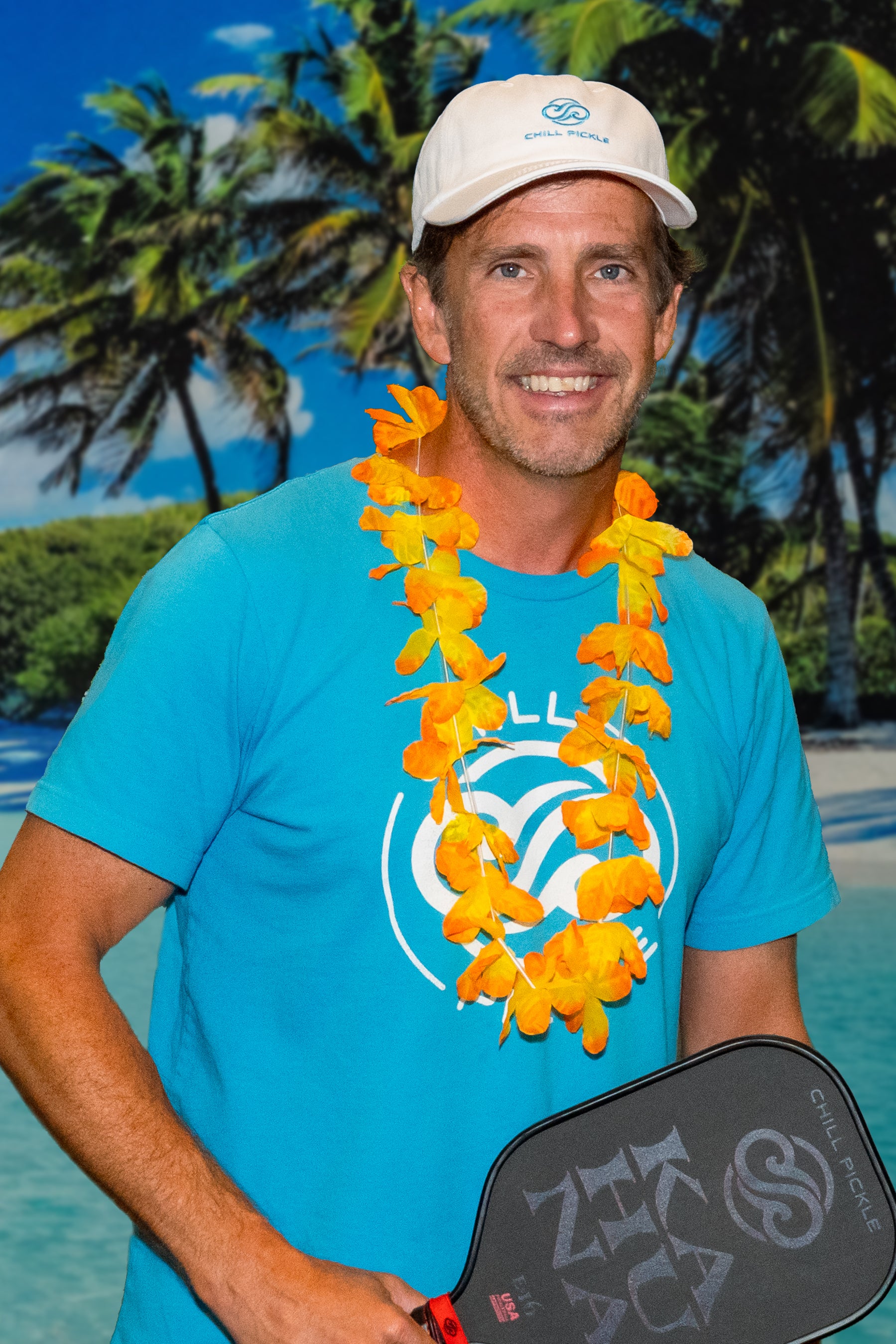 Man wearing a blue Chill Pickle t-shirt holding a pickleball paddle in a tropical outdoor setting