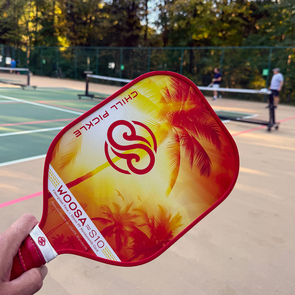 Hand holding up Chill Pickle Woosa pickleball paddle r on an outdoor court