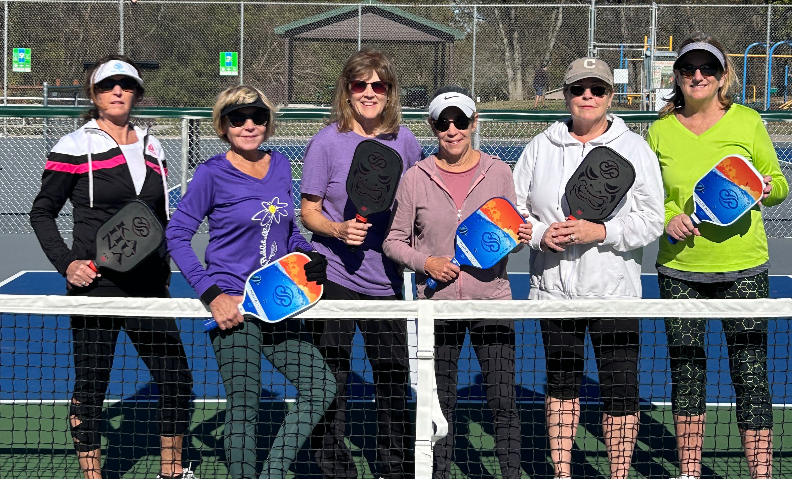 Group of women standing together on an outdoor pickleball court holding Chill Pickle paddles before a casual group match